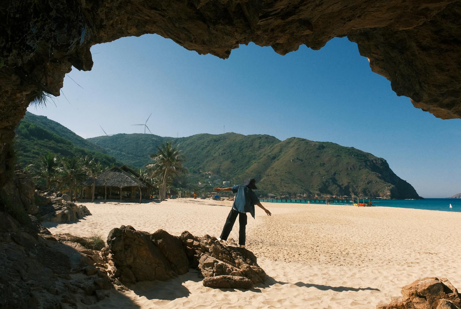 Person exploring a sandy beach with mountains and wind turbines in the background.