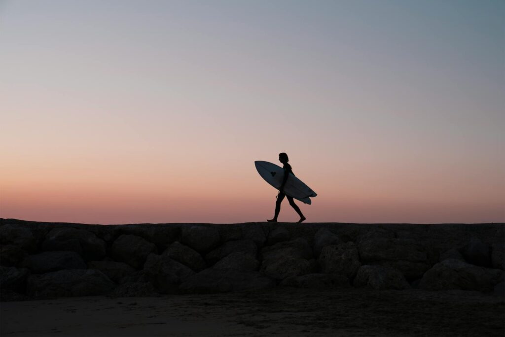 Silhouette of a surfer walking with a surfboard at sunset on a rocky beach in Portugal.