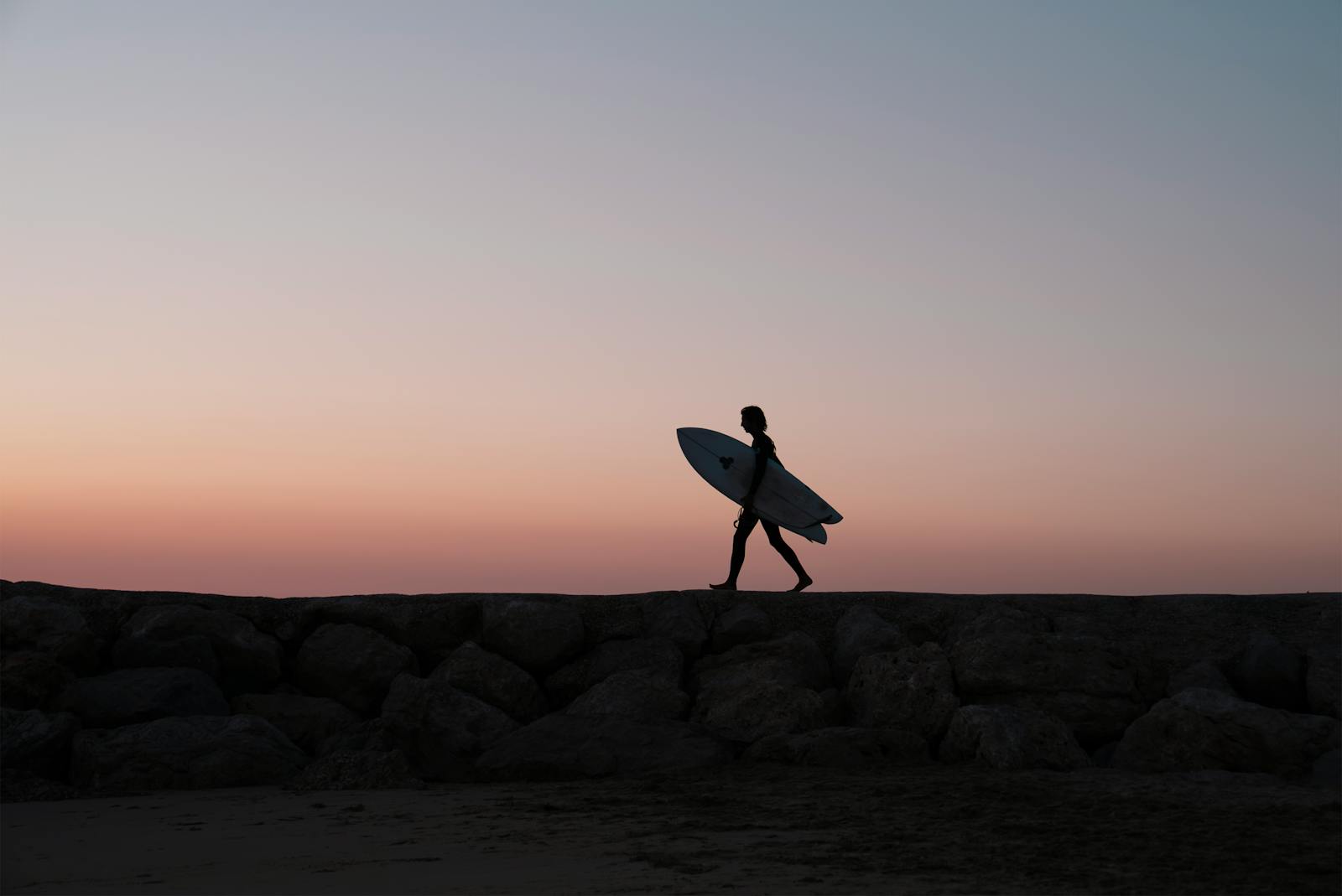 Silhouette of a surfer walking with a surfboard at sunset on a rocky beach in Portugal.