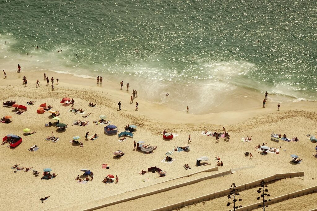 Crowded beach scene with people sunbathing and swimming on a sunny day, seen from above.