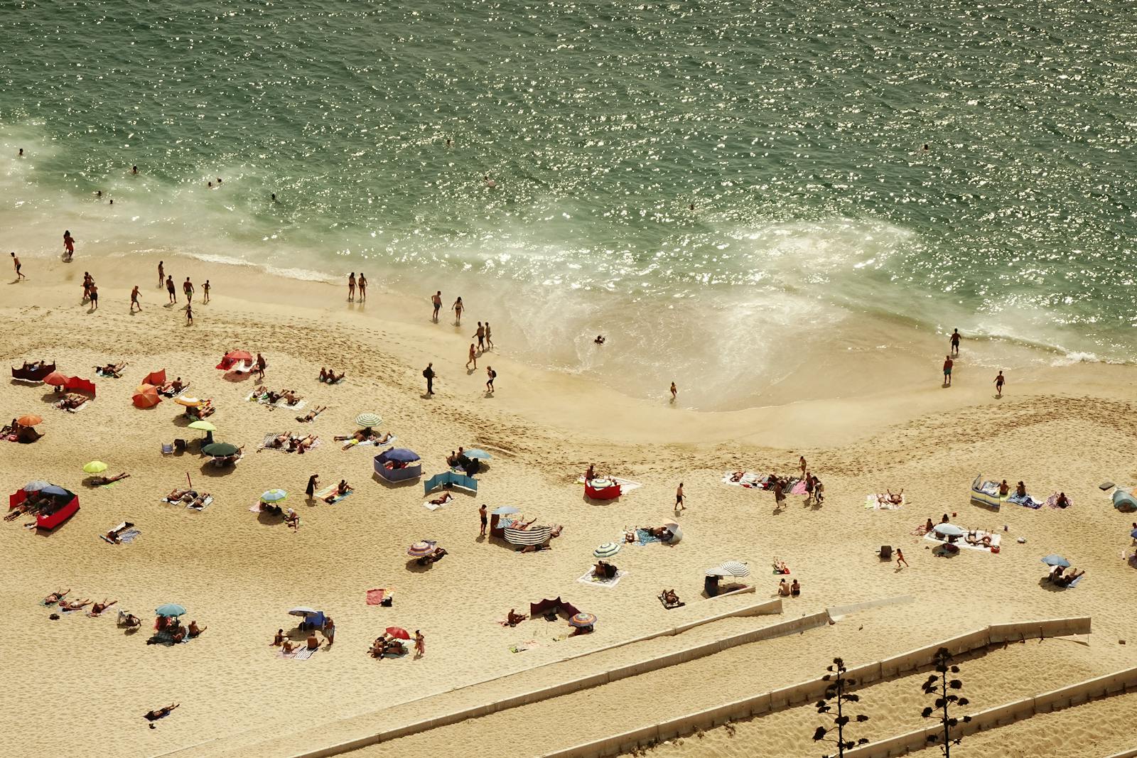 Crowded beach scene with people sunbathing and swimming on a sunny day, seen from above.